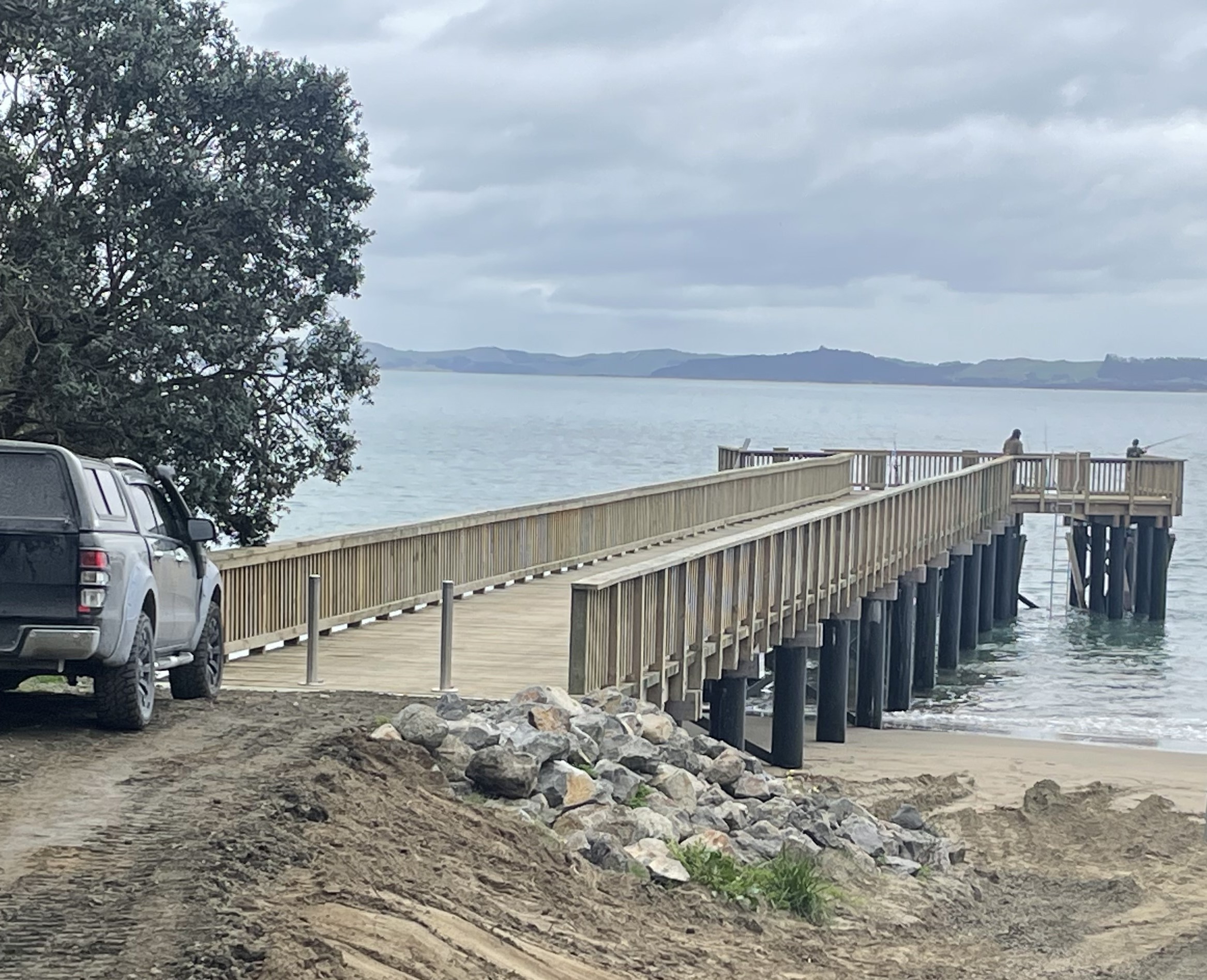 Pouto wharf stretching out into the Kaipara harbour, with two people fishing off the wharf head. 